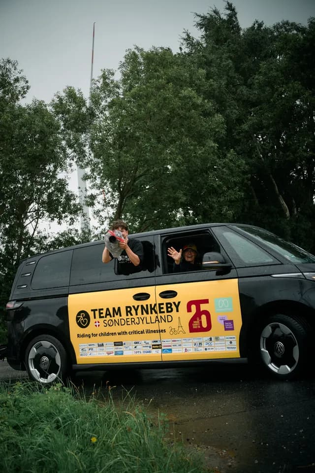 Hjælpere vinker fra en Team Rynkeby-følgebil dækket af sponsorlogoer